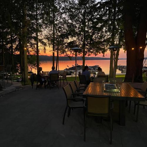 A serene lakeside scene at sunset, with people seated at a table surrounded by trees and boats in the background.