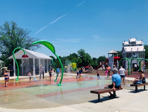 Children play in a splash pad at a park on a sunny day, with water features and playground equipment in the background.