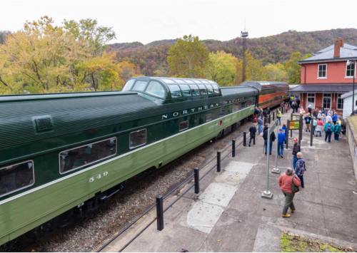 A vintage green train at a station with passengers boarding, surrounded by autumn foliage and a historic building.