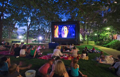 A crowd watches a movie on a large outdoor screen in a park at night, surrounded by trees and city lights.