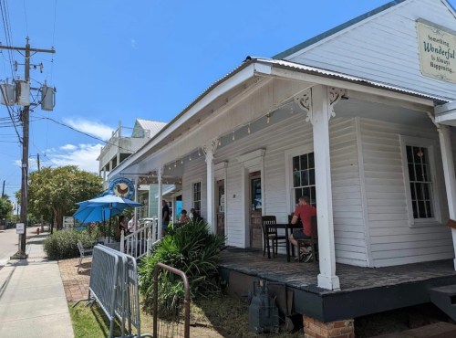 A charming white building with outdoor seating, surrounded by greenery and blue skies, inviting patrons to enjoy their meals.