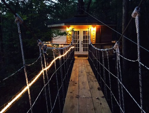 A wooden bridge with rope railings leads to a cozy treehouse illuminated by warm lights in a forest setting.