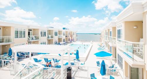 A bright pool area surrounded by modern buildings, with lounge chairs and umbrellas under a clear blue sky.