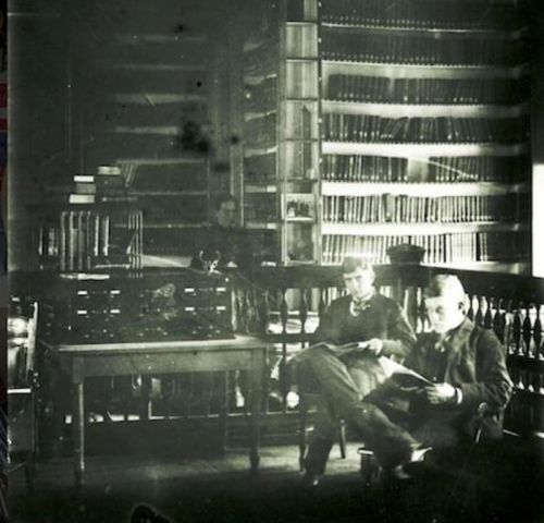 Two men reading in a dimly lit library filled with bookshelves, with a third person visible in the background.