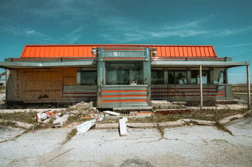 Abandoned diner with boarded windows and overgrown surroundings, featuring a bright orange roof against a clear blue sky.