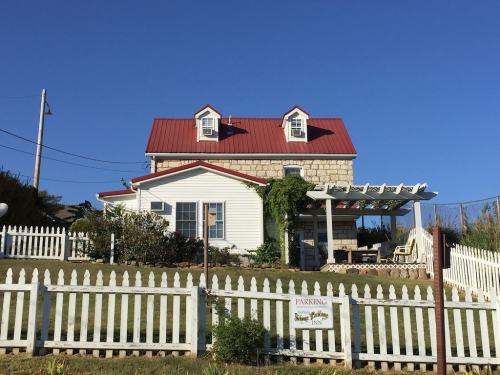 A two-story house with a red roof, white siding, and a picket fence, surrounded by greenery under a clear blue sky.