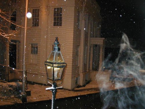 A snowy night scene featuring a vintage streetlamp and a white building, with snowflakes gently falling.
