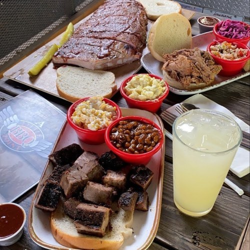 A spread of BBQ featuring ribs, pulled pork, sides like coleslaw and macaroni, and a refreshing drink on a wooden table.