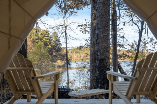 View from a tent showing two wooden chairs overlooking a serene lake surrounded by trees in autumn.