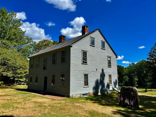 A historic gray house with a sloped roof, surrounded by green trees and a blue sky with fluffy clouds.