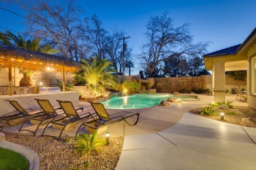 A serene backyard with a pool, lounge chairs, palm trees, and a tiki bar, illuminated at dusk.