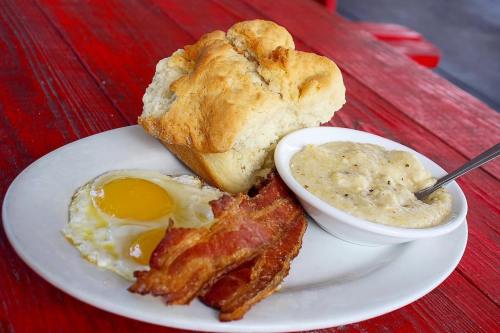 A plate with two fried eggs, crispy bacon, creamy grits, and a piece of freshly baked bread on a red wooden table.
