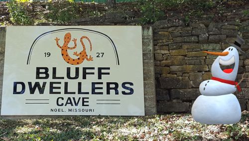 Sign for Bluff Dwellers Cave in Noel, Missouri, with a cartoon snowman beside it.
