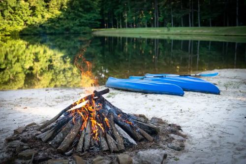 A campfire burns on a sandy shore beside a calm lake, with blue kayaks resting nearby and trees reflecting in the water.