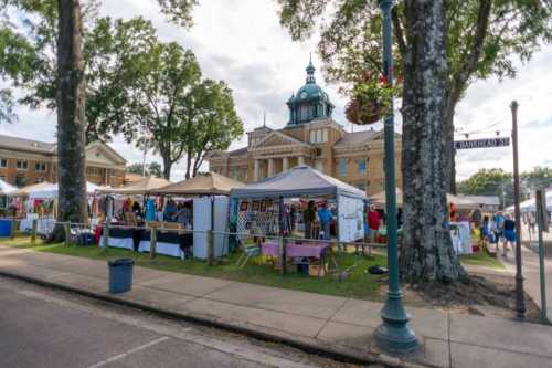 A vibrant outdoor market with tents and vendors, set against a historic building and trees.