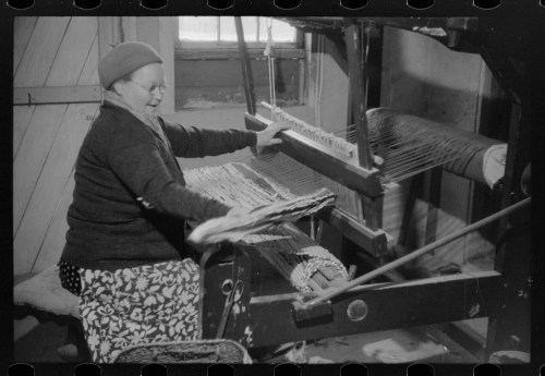 A woman weaves on a loom, smiling as she works with fabric in a cozy, rustic setting.