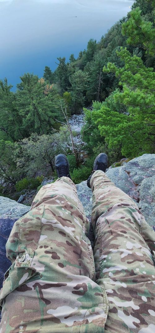 A person in camouflage pants sits on a rocky ledge, overlooking a forested area and a body of water below.