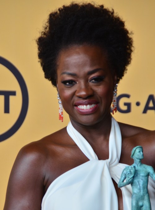 A smiling woman with curly hair holds a statue, wearing a white dress and statement earrings against a yellow background.