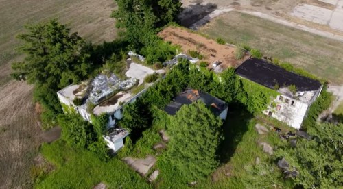 Aerial view of an overgrown, abandoned building surrounded by trees and open fields.
