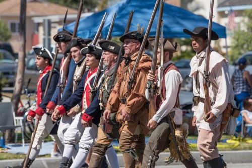 A group of historical reenactors in period costumes march with rifles during a parade.