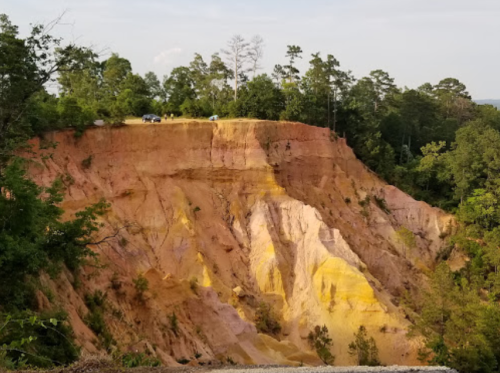 A colorful, layered cliff with pink, yellow, and orange hues, surrounded by green trees under a clear sky.