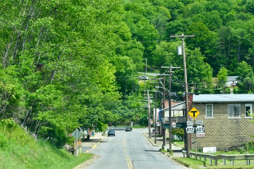 A winding road lined with trees, leading to a small town with buildings and power lines under a clear sky.