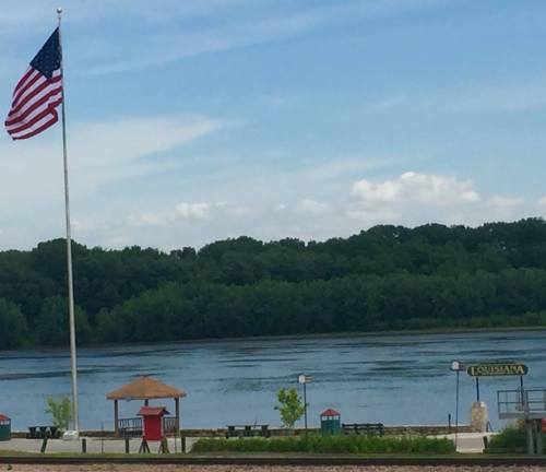 A river scene with a flagpole, American flag, and a sign for Louisiana, surrounded by greenery and a clear blue sky.