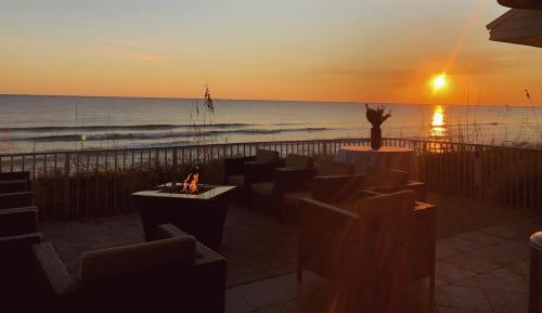 A serene beachside sunset with a fire pit and seating, casting warm hues over the calm ocean waves.