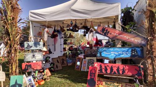 A colorful market stall with various handmade crafts, signs, and decorations, set up under a white tent.