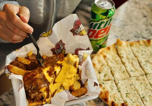 A hand holding a fork over a tray of cheesy fries with gravy, alongside a Mountain Dew and a flatbread.