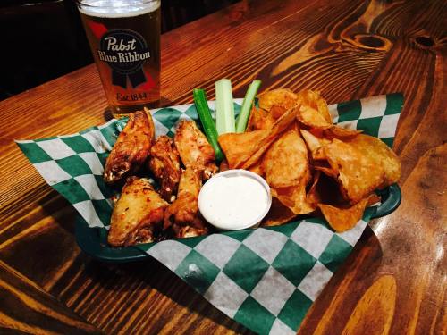 A plate of chicken wings with celery, chips, and a dipping sauce, alongside a glass of Pabst Blue Ribbon beer.