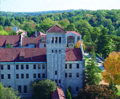 Aerial view of a historic stone building with a red-tiled roof, surrounded by trees in vibrant autumn colors.