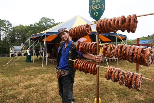 A vendor in a pirate costume stands beside a display of pretzels at a fair, with a colorful tent in the background.