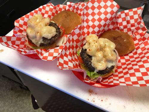 Two burgers on red and white checkered paper, topped with creamy sauce, lettuce, and tomato, with buns on the side.
