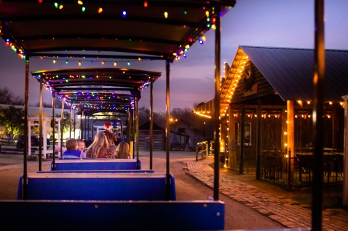 A colorful train ride at dusk, with festive lights illuminating the path and nearby buildings. Families enjoy the view.