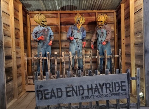 Three figures in pumpkin masks and overalls stand behind a wooden fence in a spooky, rustic setting.