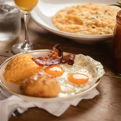 A plate with two fried eggs, bacon, a biscuit, and a side of creamy grits, with a drink in the background.