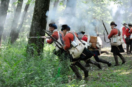 Reenactors in red coats advance through a smoky forest, carrying muskets and wearing historical military gear.