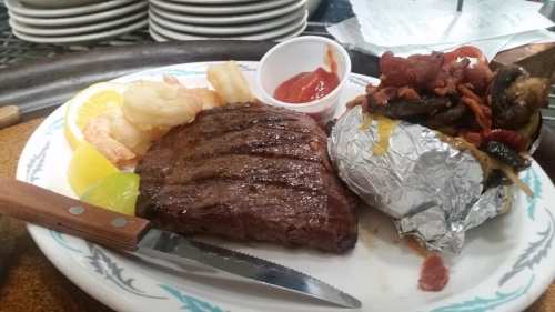 A plate featuring grilled steak, shrimp, a baked potato with toppings, and dipping sauce, garnished with lemon and lime.