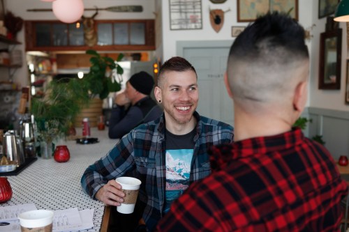 Two people chatting at a café, one smiling and holding a coffee cup, with a cozy atmosphere and plants in the background.