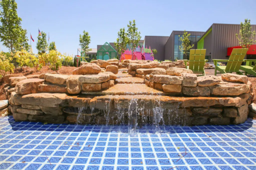 A stone water feature with cascading water over rocks, surrounded by greenery and colorful buildings in the background.