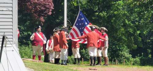 A group of people in colonial attire is raising an American flag outdoors, surrounded by greenery.