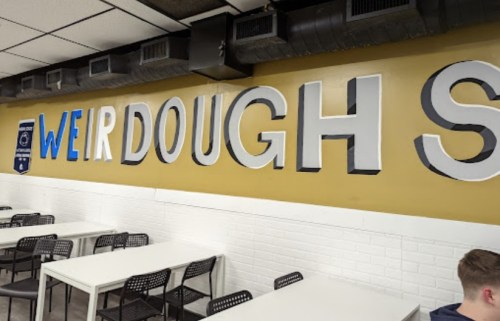 A wall with the words "WEIRD DOUGH" in large, colorful letters above empty tables and chairs in a casual dining space.