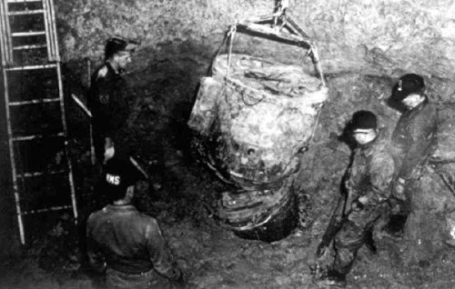 A group of men examines a large, suspended object in a pit, with a ladder visible in the background.