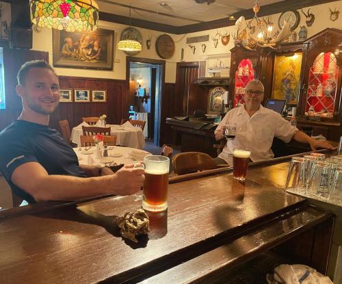 Two men sit at a bar, smiling and holding large beers, with a cozy, rustic restaurant atmosphere in the background.