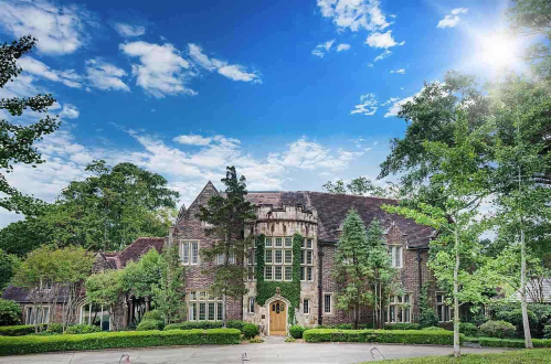 A large, elegant stone house surrounded by lush greenery and trees under a bright blue sky with scattered clouds.