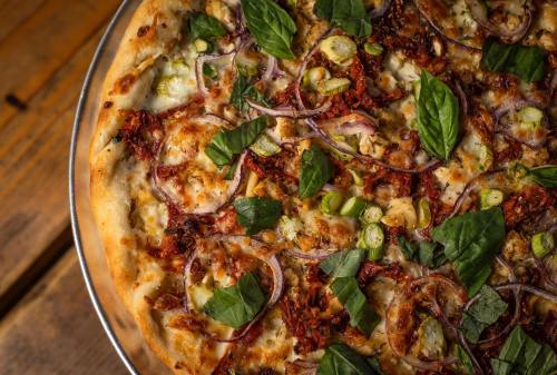 A close-up of a pizza topped with sun-dried tomatoes, red onions, green onions, and fresh basil on a wooden table.