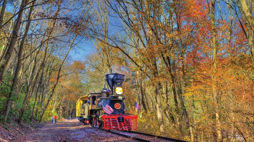 A vintage steam train travels along a scenic track surrounded by colorful autumn trees.