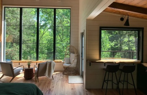 Cozy interior with large windows overlooking greenery, featuring a seating area and a small kitchen bar.