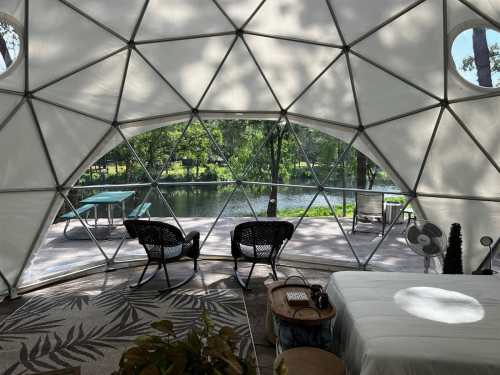 Interior of a geodesic dome with two chairs, a bed, and a view of a serene lake and greenery outside.